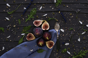 From above fresh halved figs and silver spoon placed on wooden table near blue napkin and arugula in kitchen