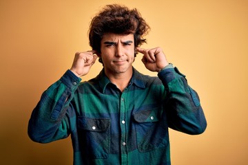 Young handsome man wearing casual shirt standing over isolated yellow background covering ears with fingers with annoyed expression for the noise of loud music. Deaf concept.