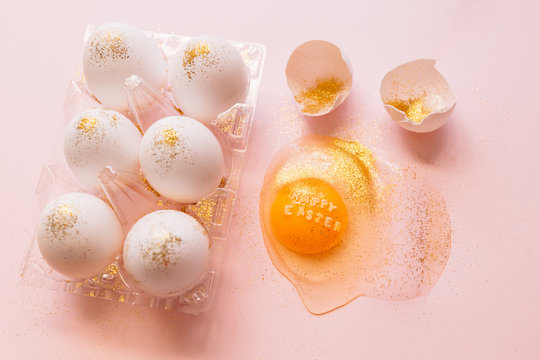 Top view of broken glittered egg with text Happy Easter and raw eggs in plastic box on pink background