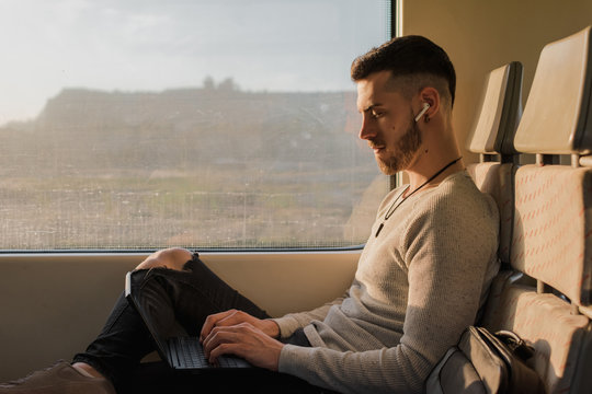Side view of young man in wireless earphones typing on laptop during journey in subway in sunny day