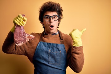 Young handsome butcher man holding meet steak standing over isolated yellow background Surprised...