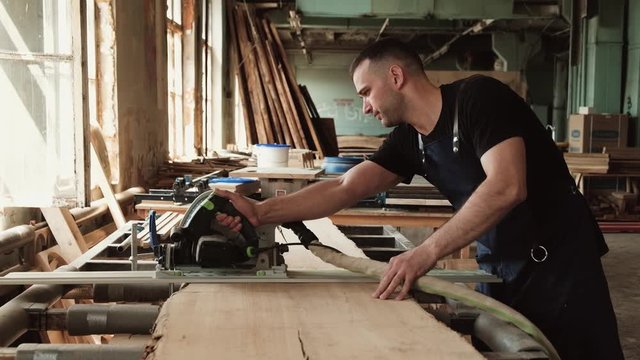 young male carpenter cutting wood board with a saw. Joiner sawing a big piece of wood in industrial workshop. self-employed man and his small business.