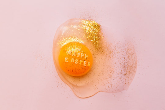Top view of broken raw egg with text Happy Easter on yolk covered with golden sparkles on pink background