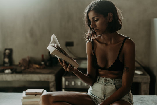 Interested Youthful Ethnic Woman In Bra And Short Enjoying Reading A Book While Sitting On Marble Countertop In Kitchen