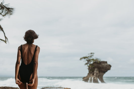 Back view of woman in black swimsuit walking on empty sandy coast with stormy ocean in overcast on background