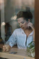 Beautiful young woman sits in a cafe behind a glass with a cup of coffee and and looks into a smartphone. Photo through glass with reflection. Soft selective focus.