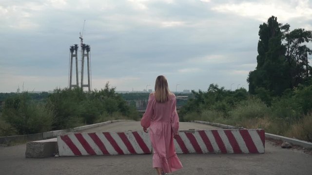 Overall plan. beautiful, young girl with long hair pink dress walks alone along asphalt road, past concrete red blocks, turns sharply. Against background unfinished bridges, lampposts, metal
