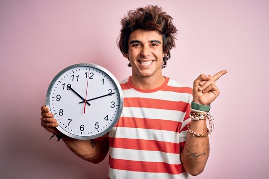 Young handsome man holding big clock standing over isolated pink background very happy pointing with hand and finger to the side