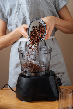 Crop female in grey apron putting soaked lentils from bowl to food processor while preparing dinner at wooden table in kitchen