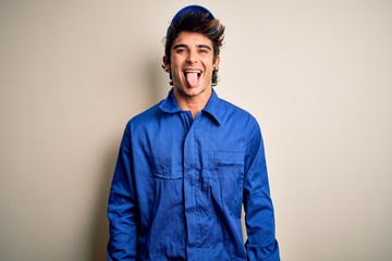Young mechanic man wearing blue cap and uniform standing over isolated white background sticking tongue out happy with funny expression. Emotion concept.