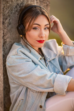 Side view of young woman looking at camera in casual wear resting on rocky fence listening music with headphones