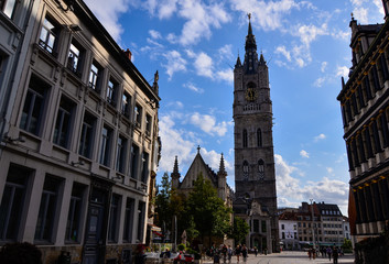 Fototapeta na wymiar Ghent, Belgium, August 2019. The imposing bell tower called Beffroi, with its grandeur stands out compared to the surrounding houses. People on street. Sunny day.