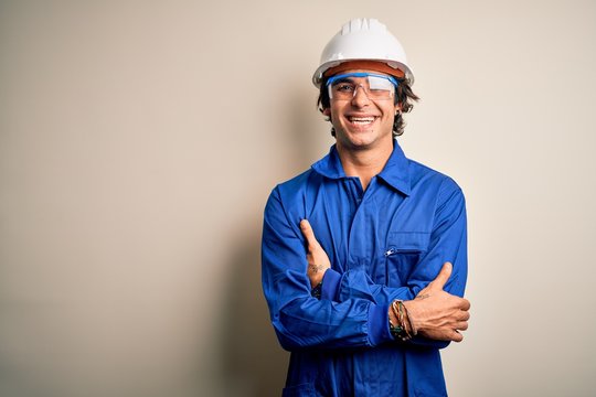 Young constructor man wearing uniform and security helmet over isolated white background happy face smiling with crossed arms looking at the camera. Positive person.