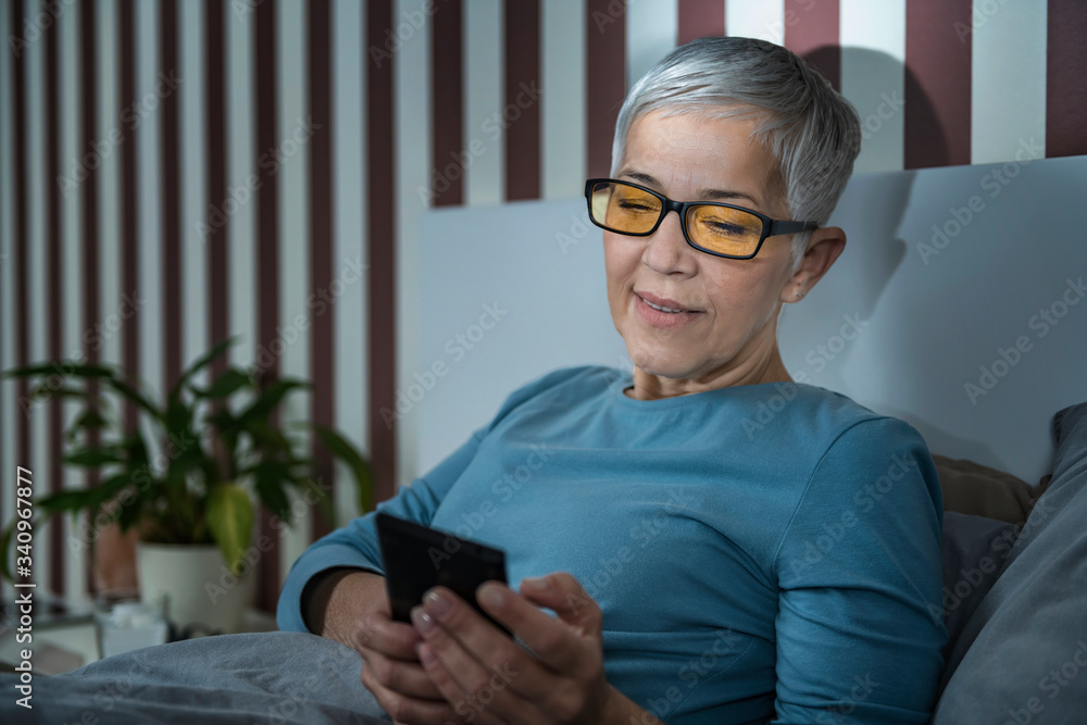 Wall mural Senior Woman with Blue Light Blocking Glasses Using Smartphone in the Evening