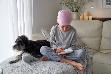 From below happy little child with cancer disease writing notes while sitting with dog on bed in room