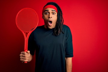 African american afro sportsman with dreadlocks holding tennis racket over red background scared in shock with a surprise face, afraid and excited with fear expression