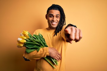 Young african american afro romantic man with dreadlocks holding bouquet of yellow tulips pointing to you and the camera with fingers, smiling positive and cheerful