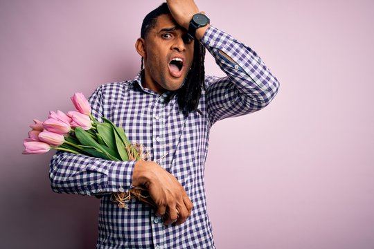 Young african american afro romantic man with dreadlocks holding bouquet of pink tulips surprised with hand on head for mistake, remember error. Forgot, bad memory concept.
