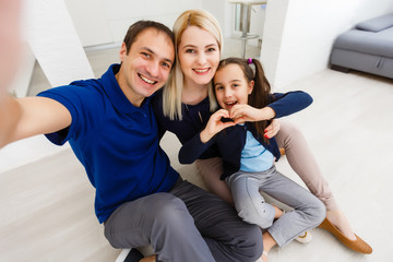 Beautiful young mother, father and their daughter are making selfie using a phone and smiling while sitting at home