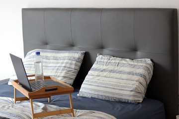 a wooden serving tray with a computer, a bottle of water and a red handle stands on a bed in blue linen