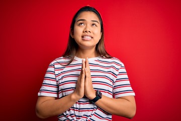 Young beautiful asian girl wearing casual striped t-shirt over isolated red background begging and...