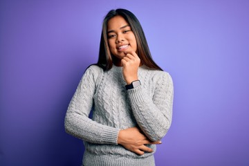 Young beautiful asian girl wearing casual sweater standing over isolated purple background looking confident at the camera with smile with crossed arms and hand raised on chin. Thinking positive.