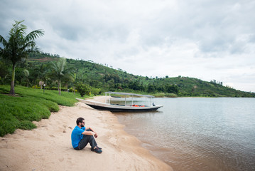 Fototapeta na wymiar Caucasian male tourist sitting on Lake Kivu shore observing the water with green lush vegetation in the background