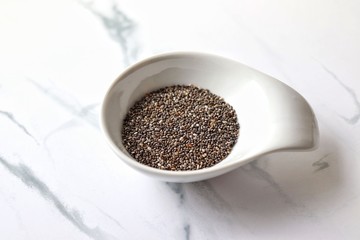 Closeup of chia seeds in white dish, on marble table. Macro, horizontal.