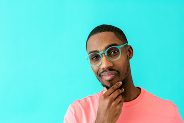 Obraz premium Portrait of a young man with glasses thinking, deciding and holding his chin, against blue studio background