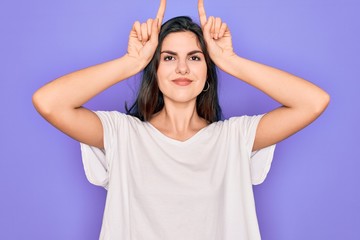 Obraz na płótnie Canvas Young beautiful brunette woman wearing casual white t-shirt over purple background doing funny gesture with finger over head as bull horns