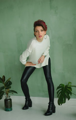 Full height studio portrait of tall young fashion model wearing white sweater and black leather leggings possing at studio with green background and exotic plants