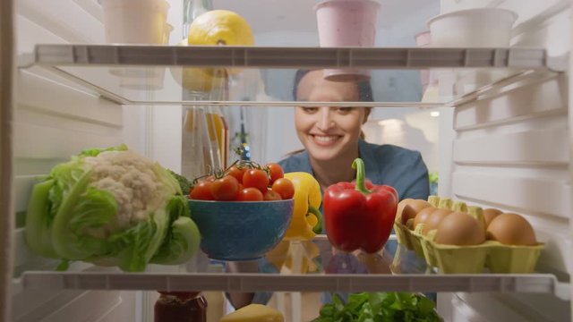 Beautiful Young Woman Opens Fridge Door, Looks inside Takes out Vegetables. Woman Preparing Healthy Meal Using Groceries. Point of View POV from Inside of the Kitchen Refrigerator full of Healthy Food