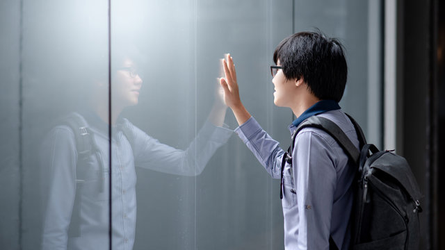 Young Asian Man Carrying Bag Looking At Himself Touching On Glass Window While Raining. Guy Reflecting In The Mirror. Emotional Reflection Concept