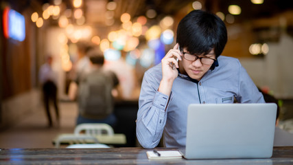 Smart Asian businessman wearing glasses working with laptop computer and smartphone. Male entrepreneur using phone for business talk. Technology for business management and strategy.