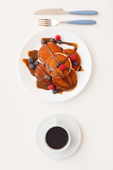 Top view at minimal composition of delicious golden pancakes decorated with chocolate and fresh berries next to coffee cup on white background, breakfast and dessert concept, copy space