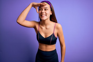 Young beautiful sporty girl doing sport wearing sportswear over isolated purple background very happy and smiling looking far away with hand over head. Searching concept.