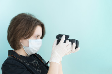 young female photographer in medical mask and white medical gloves holds her camera on light blue backgrouund, protection from viruses and coronavirus pandemic, health care concept