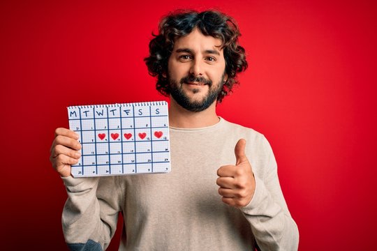 Young handsome man with beard holding calendar over isolated red background happy with big smile doing ok sign, thumb up with fingers, excellent sign