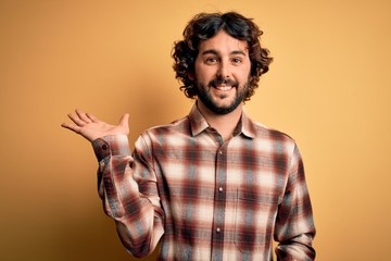 Young handsome man with beard wearing casual shirt standing over yellow background smiling cheerful presenting and pointing with palm of hand looking at the camera.