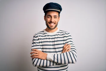 Poster Young handsome sailor man with beard wearing navy striped uniform and captain hat happy face smiling with crossed arms looking at the camera. Positive person. © Krakenimages.com