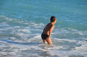 A small, happy boy stands in the water and splashes. A cheerful child jumps on the sea shore, the beach. The child enjoys the sea and splashes in the water with cottage cheese. Rear view.