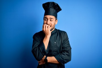 Young handsome chef man with beard wearing cooker uniform and hat over blue background looking stressed and nervous with hands on mouth biting nails. Anxiety problem.