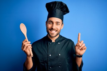 Fototapeta premium Young cooker man with beard wearing uniform holding wooden spoon over blue background surprised with an idea or question pointing finger with happy face, number one