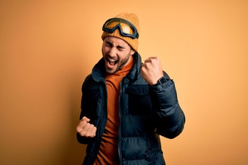 Young handsome skier man with beard wearing snow sportswear and ski goggles celebrating surprised...