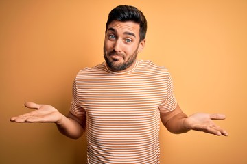 Young handsome man with beard wearing casual striped t-shirt over yellow background clueless and...