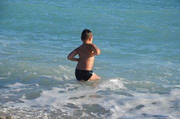 A small, happy boy stands in the water and splashes. A cheerful child jumps on the sea shore, the beach. The child enjoys the sea and splashes in the water with cottage cheese. Rear view.