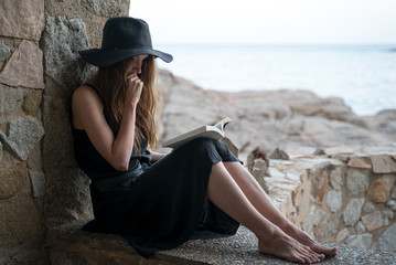 Girl reads book. A girl in a black hat and black silk dress sits in the niche of a fishing hut on the seashore and reads a book. Ocean view at a background. Studying outdoors, reading a novel.