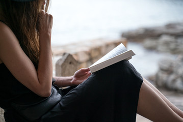 Girl reads book. A girl in a black hat and black silk dress sits in the niche of a fishing hut on the seashore and reads a book. Ocean view at a background. Studying outdoors, reading a novel.