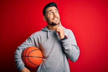 Young sports man holding basketball ball over red isolated background serious face thinking about question, very confused idea