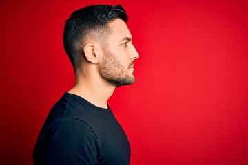 Young handsome man wearing casual black t-shirt standing over isolated red background looking to...